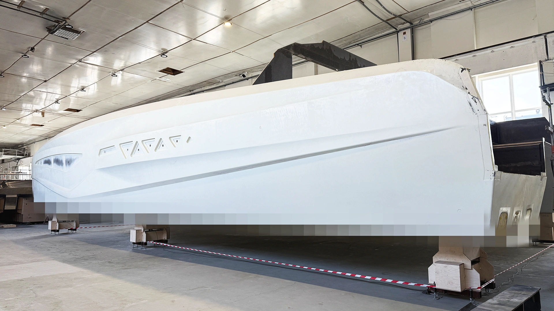 Side view of a white, unfinished boat hull with geometric window cutouts, supported by wooden blocks in a manufacturing facility
