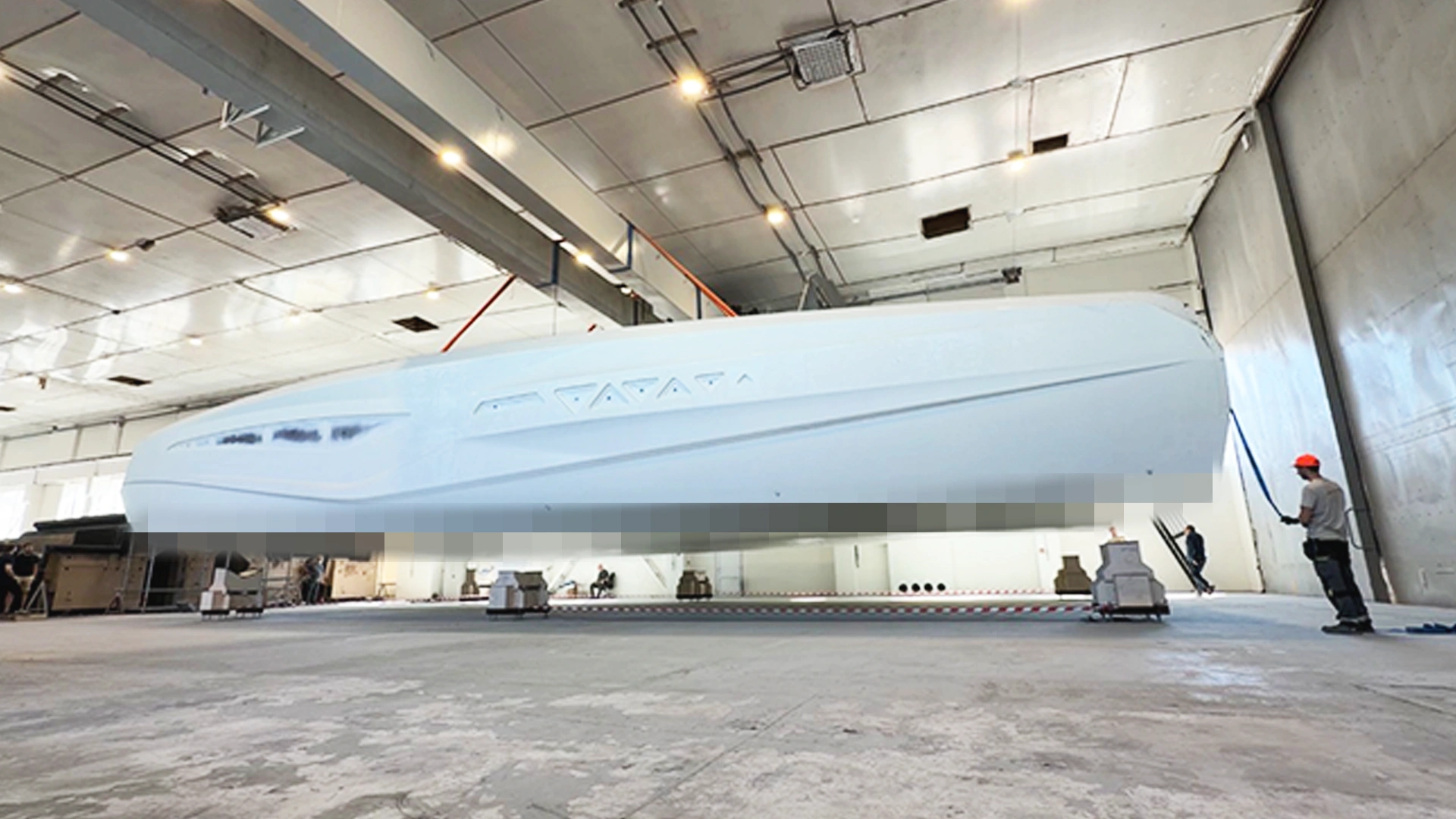 A large white boat hull suspended in the air inside a warehouse, with a worker in an orange hard hat standing nearby